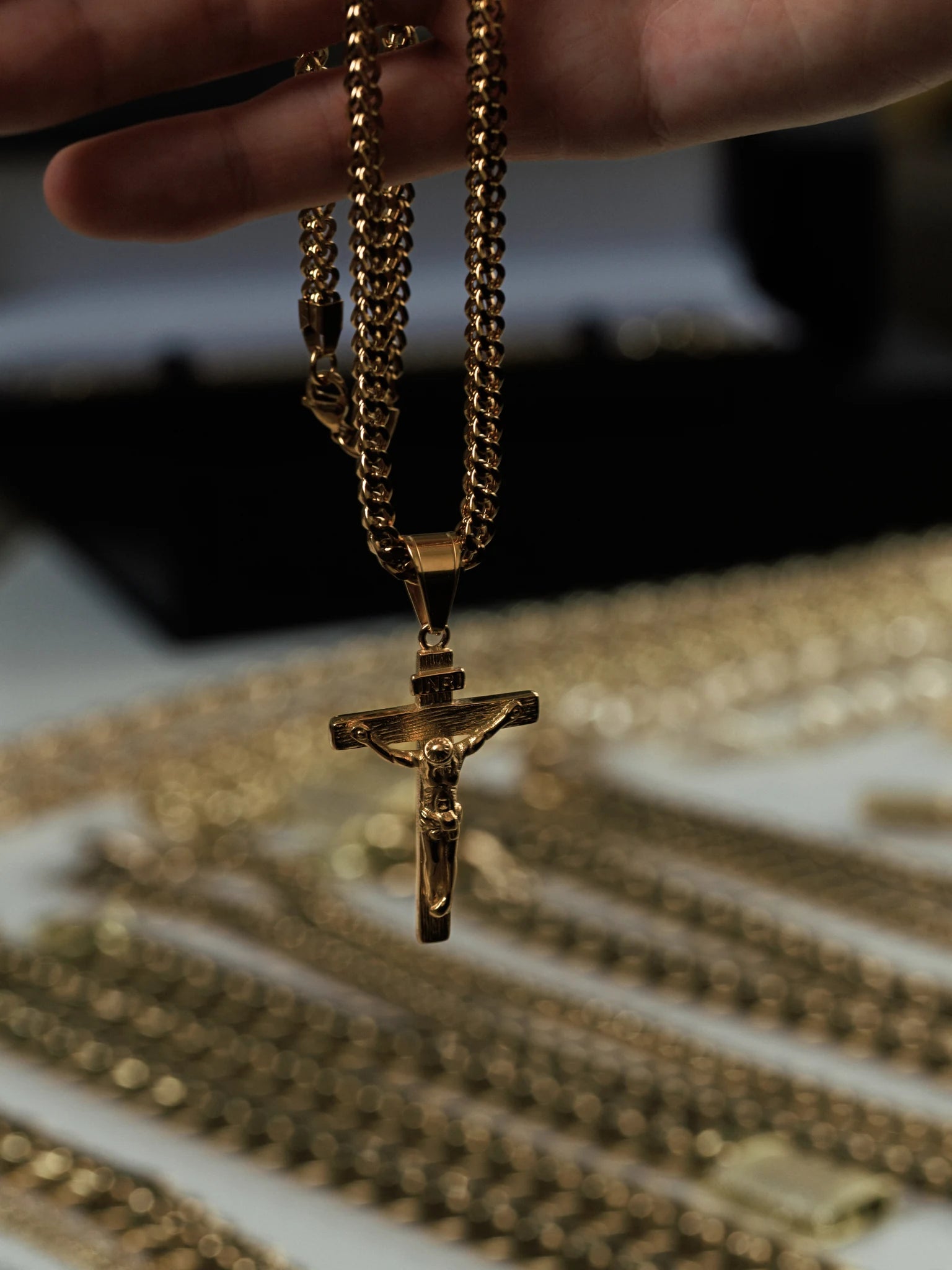Gold filled crucifix pendant on a 4mm Franco chain held in hand above a tray of gold chains